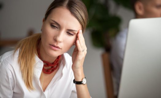 A woman in a white shirt looking stressed while sitting at her desk with a computer, touching her temple.