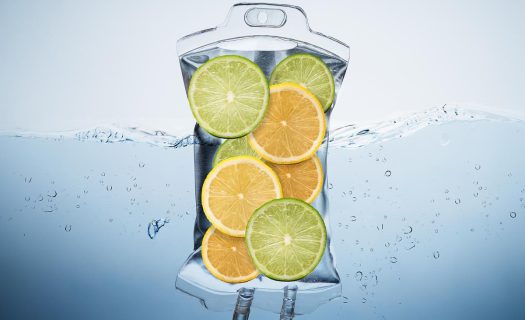 Transparent water pitcher filled with orange and lime slices, partially submerged in water with bubbles around it.