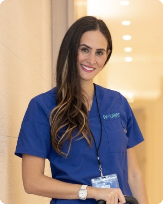 A female healthcare professional in blue scrubs and an id badge smiles while standing in a hospital corridor.