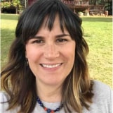 A smiling woman with shoulder-length brown hair and bangs, wearing a grey sweater and a blue necklace, outdoors with trees in the background.