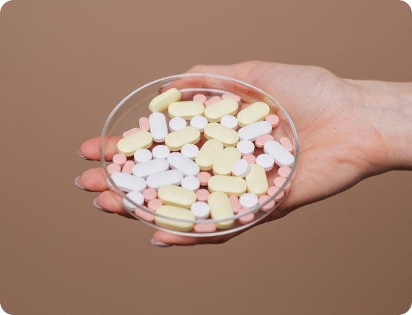 A hand holding a clear dish filled with various colored pills against a neutral background.