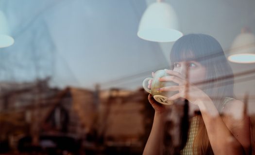 A woman drinking coffee at a cafe table, viewed through a window with reflections. she has bangs and is holding a large cup with both hands.