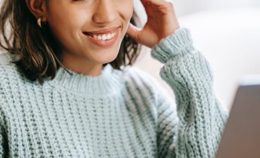 Young woman smiling and looking at her laptop screen, wearing a teal sweater, with her hand resting on her head.