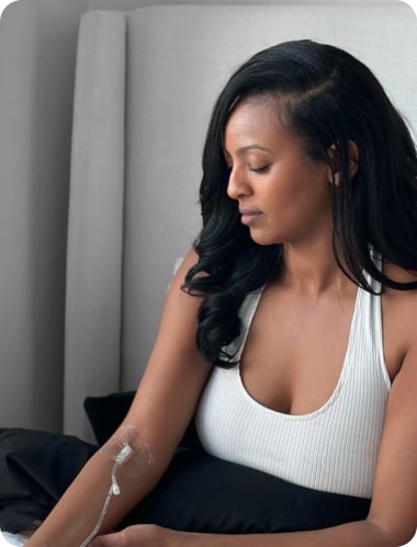 A woman with an iv line in her arm sits pensively on a gray couch, wearing a white tank top, looking downward.