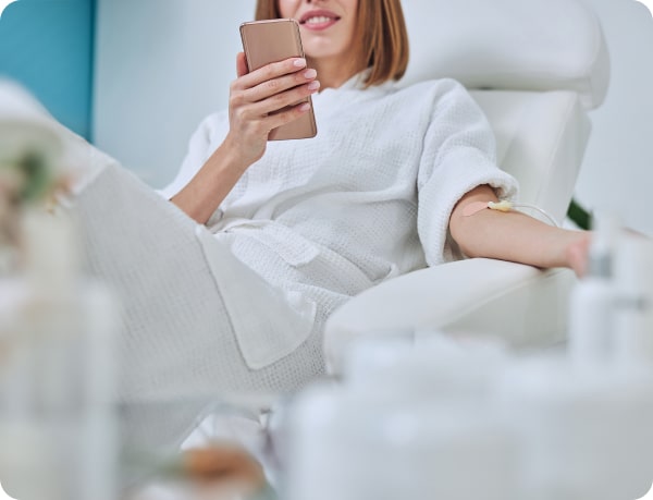 Woman in a white robe relaxing on a chair, using a smartphone, with a focus on comfort and modern technology in a cozy setting.