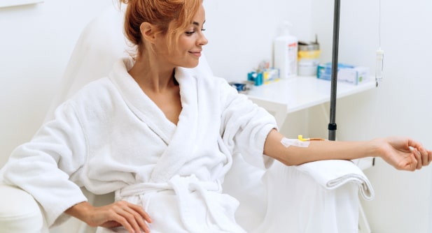 A woman in a white robe smiles while receiving an intravenous treatment in a serene medical setting.