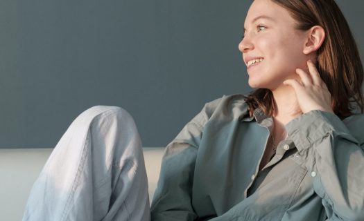 Young woman in a blue shirt sitting and smiling while looking to the side, hand on chin, against a gray background.