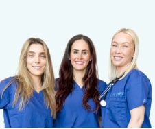 Three female healthcare professionals in blue scrubs standing together and smiling against a white background.