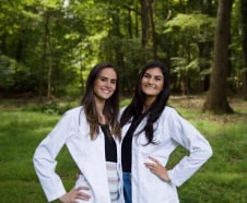 Two women in white lab coats standing outdoors with trees in the background, smiling at the camera.