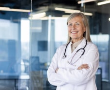 A smiling female doctor with blond hair, wearing a white coat and stethoscope, stands confidently in a modern office setting.