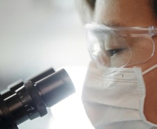 Scientist in safety goggles and face mask closely examining a specimen through a microscope in a laboratory setting.