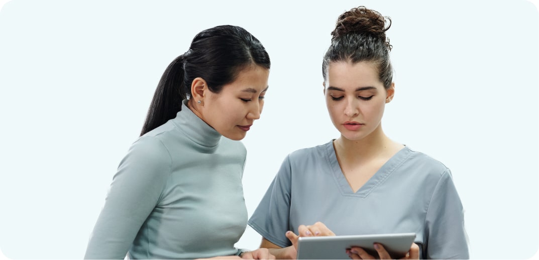 Two women, one asian and one caucasian, in casual attire, are intently looking at a tablet they are both holding.