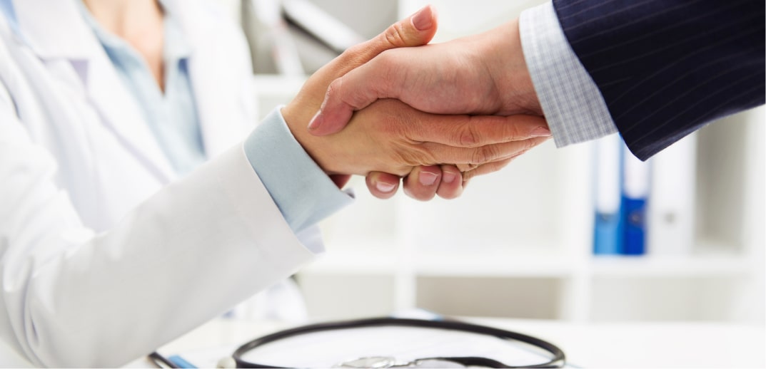Close-up of a handshake between a doctor in a white coat and a person in a business suit over a desk, with a stethoscope visible in the background.