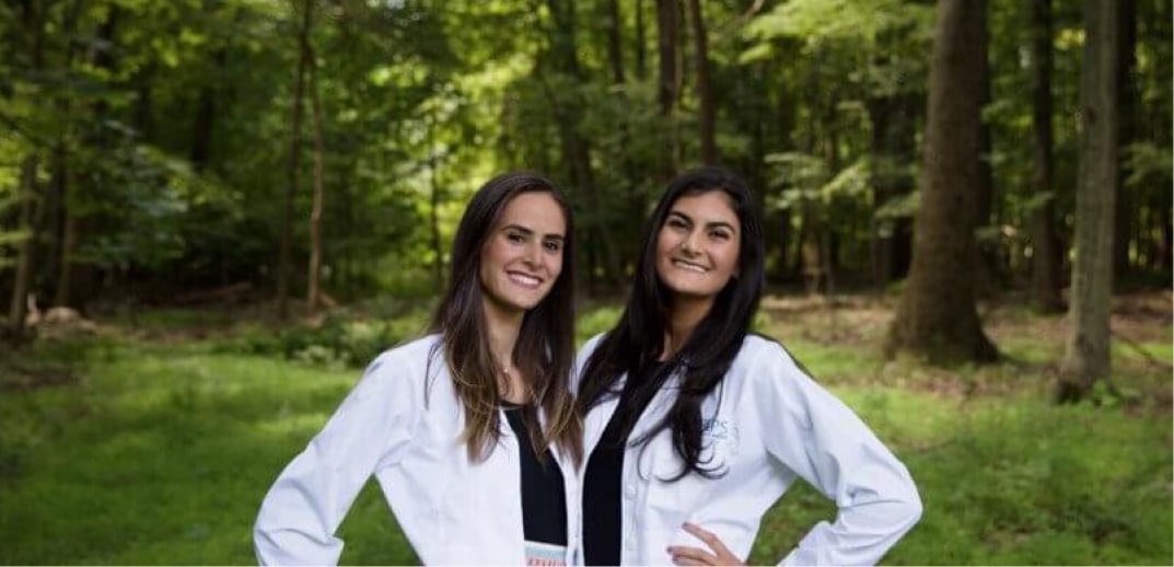 Two women in white lab coats standing confidently in a forest, smiling at the camera.