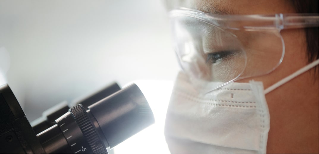 Scientist wearing safety goggles and a face mask peers through a microscope in a laboratory setting.