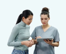 Two women, one in a medical scrub and the other in a turtleneck, looking at a tablet together against a light background.