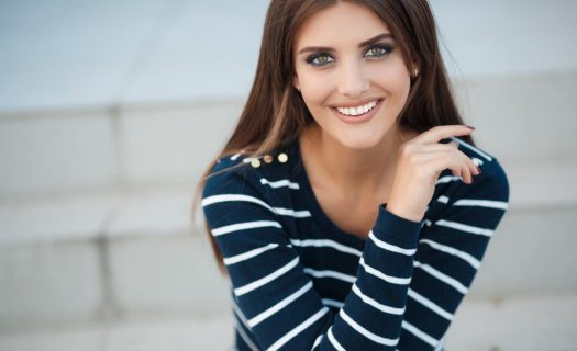 A young woman with long brown hair, smiling, wearing a striped navy and white top, sitting against a light gray wall.