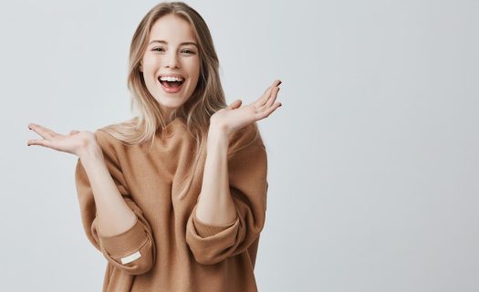 A joyful young woman in a beige sweater, with her hands raised in a welcoming gesture, smiling broadly on a light background.