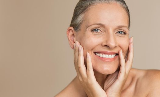 Mature woman smiling, holding her face with both hands, against a neutral background.