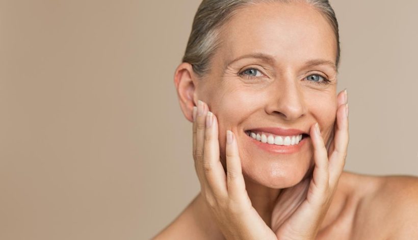 Mature woman smiling, holding her face with both hands, against a neutral background.