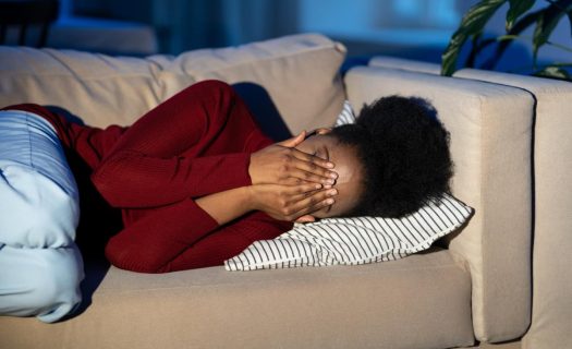 Woman lying on a couch, covering her face with her hands, appearing distressed or overwhelmed.