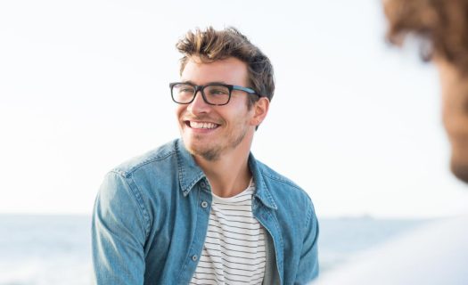 Smiling man with glasses wearing a denim jacket and striped shirt, looking at someone off-camera by the sea.