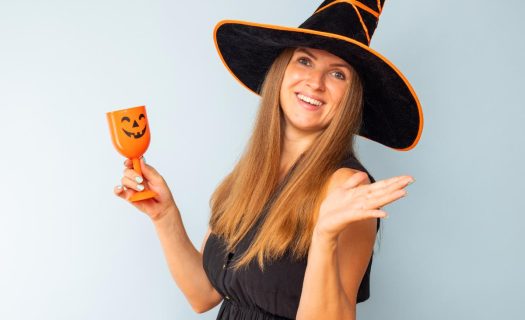 A woman in a witch hat smiling and holding an orange cup with a pumpkin face, gesturing with her hand, against a light blue background.