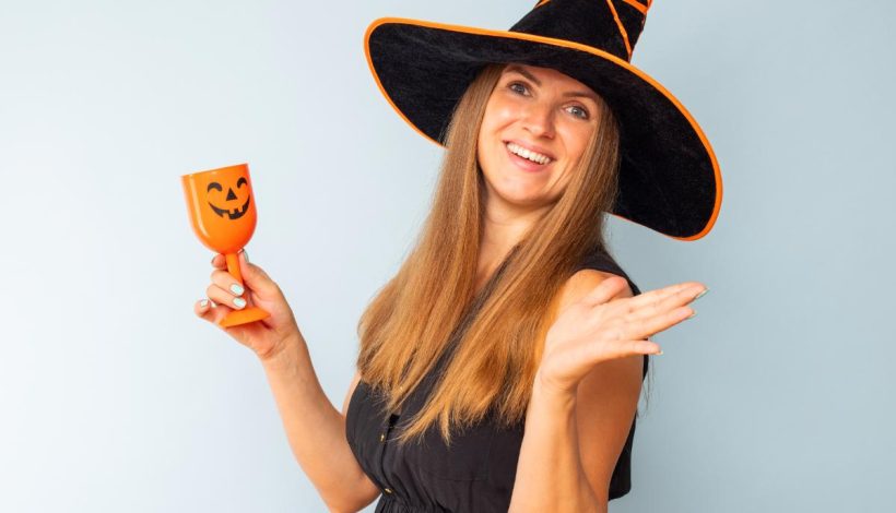 A woman in a witch hat smiling and holding an orange cup with a pumpkin face, gesturing with her hand, against a light blue background.