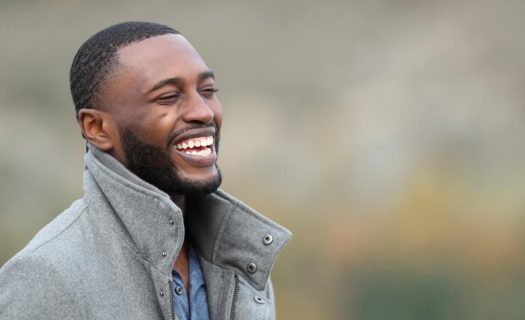 A joyful black man with a beard, wearing a gray jacket, laughing outdoors with a blurred natural background.