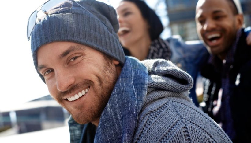 A smiling man in a beanie and scarf with friends laughing in the background.