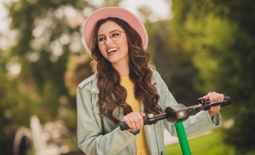 A joyful woman in a pink hat and glasses rides an electric scooter in a sunny park.