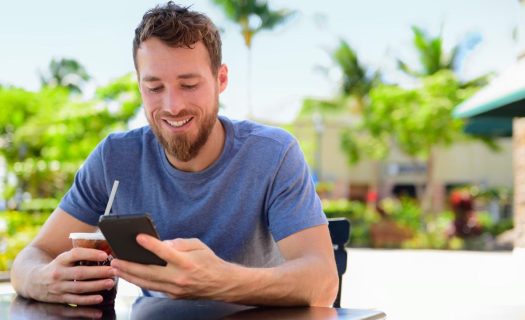 A man smiling as he uses a smartphone at a cafe table outdoors, holding a drink with a straw.