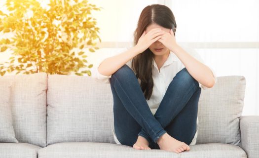 A woman sits on a couch, covering her face with her hands, appearing distressed, in a brightly lit living room.