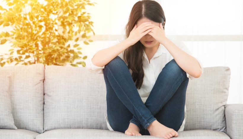 A woman sits on a couch, covering her face with her hands, appearing distressed, in a brightly lit living room.