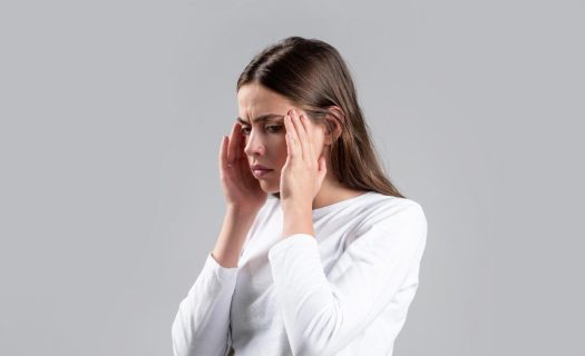 A woman in a white shirt touching her temples, looking stressed or in pain, against a gray background.