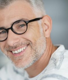 A man with gray hair and beard, wearing round glasses and a white shirt with gray stripes, smiles at the camera.