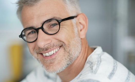 A man with gray hair and beard, wearing round glasses and a white shirt with gray stripes, smiles at the camera.