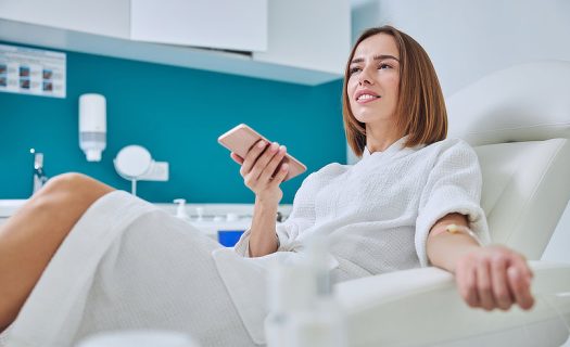 A woman in a white robe sits in a medical chair, holding a smartphone in one hand with a medical bandage on her other arm. She appears to be in a clinic or medical facility.