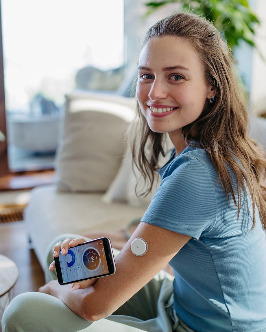 A woman with a glucose monitor on her arm holds a smartphone displaying health data, sitting on a sofa and smiling at the camera.