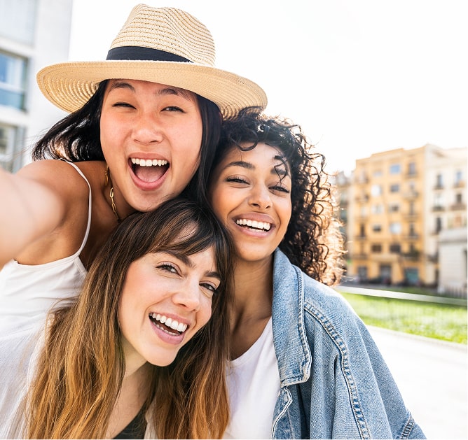 Three young women smile and pose for a selfie outdoors on a sunny day, with buildings visible in the background.