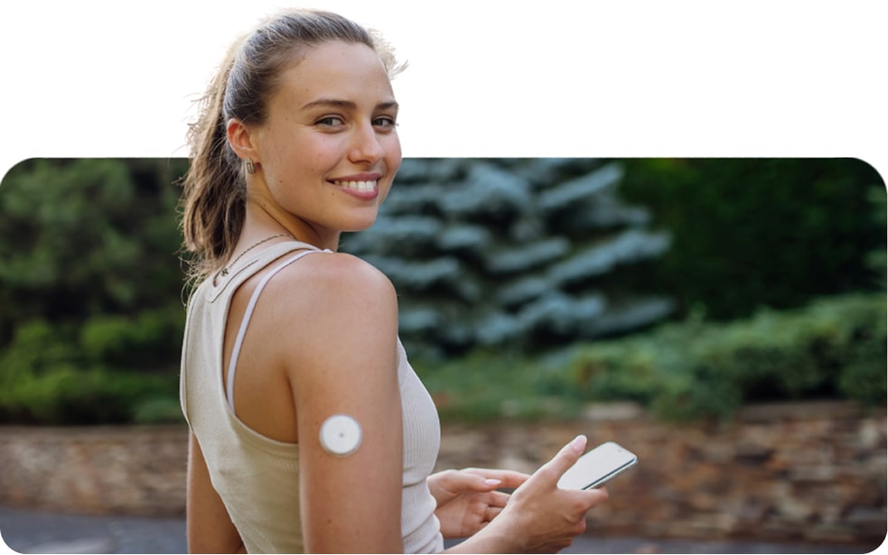 A woman outdoors smiles at the camera, holding a smartphone. She has a circular medical sensor attached to her upper arm.