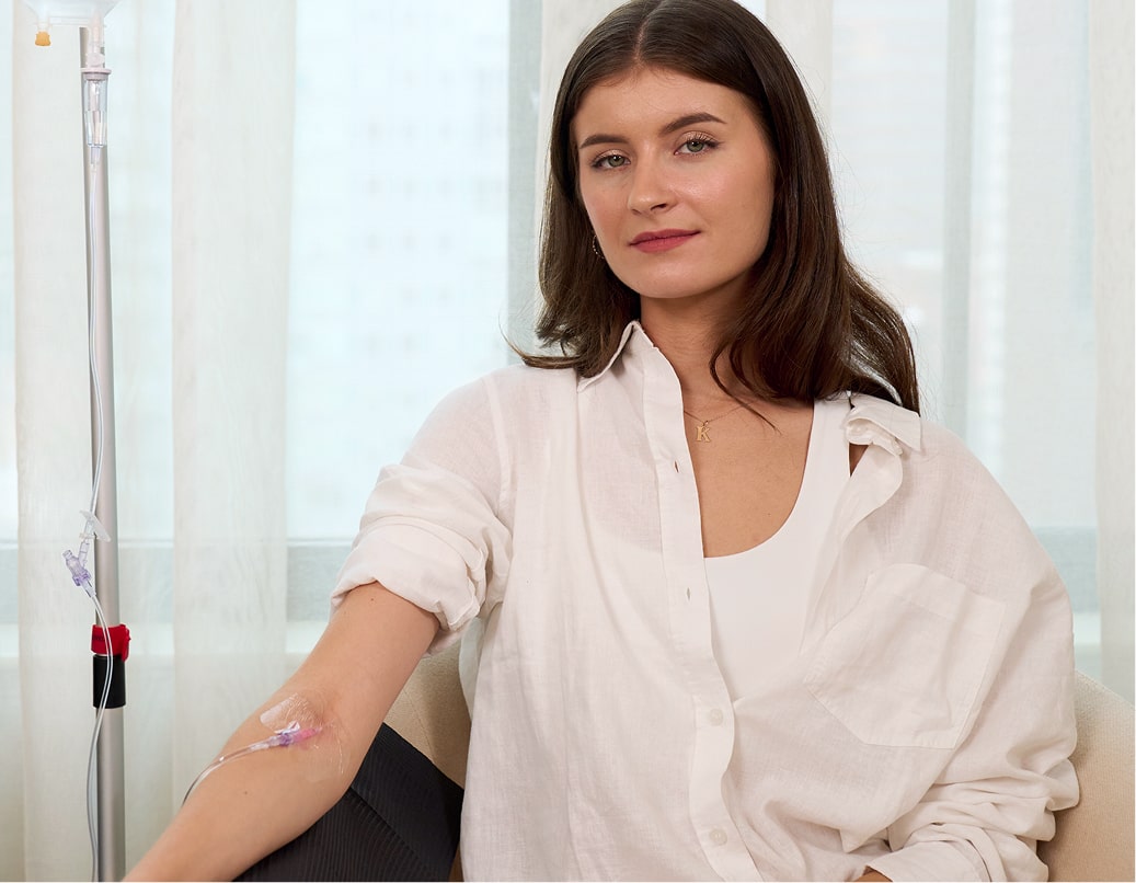 A woman with long brown hair, wearing a white shirt, sits in a chair with an IV drip attached to her arm. She looks calmly at the camera, with a window and sheer curtains in the background.