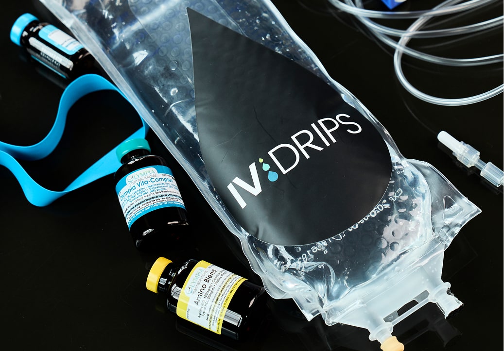 A close-up of an IV bag labeled IV Drips with medical vials, a blue tourniquet, tubing, and a syringe scattered on a black surface.