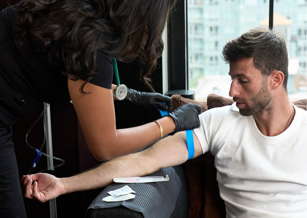 A man in a white t-shirt sits on a couch as a healthcare worker wearing black gloves prepares to administer the Recovery Drip, with medical supplies and a stethoscope nearby.