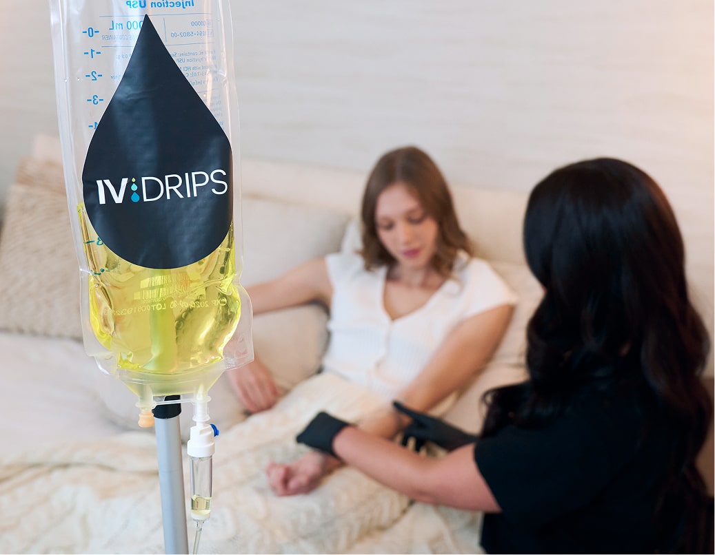 A woman sits on a bed receiving an IV drip from a healthcare professional in black gloves. The IV bag in the foreground contains a yellow liquid and is labeled Detox Drip.