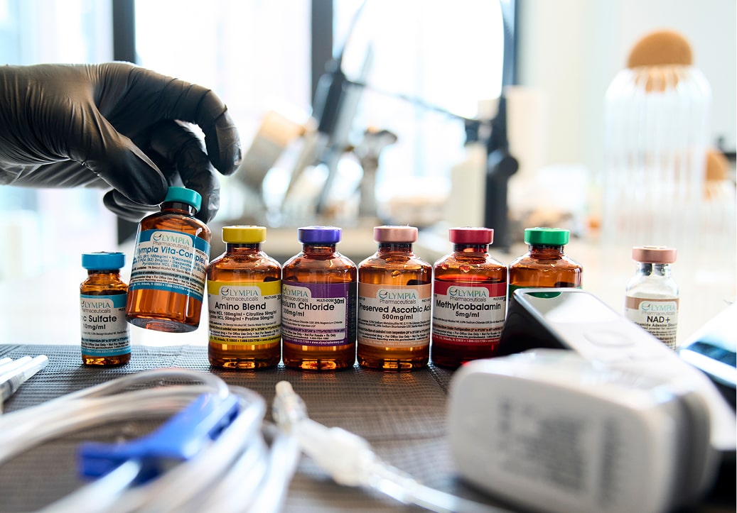 A gloved hand selects a vial labeled Custom Drip from a row of medication vials on a table, with medical equipment and a blurred laboratory background visible.
