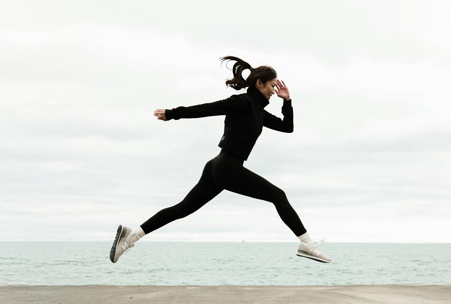 A person in black athletic clothing and white sneakers is captured mid-jump outdoors, with the ocean and a cloudy sky in the background.