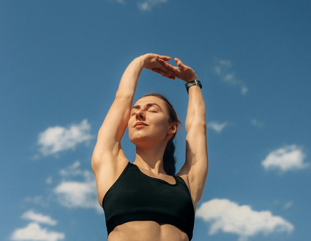 A woman in a black sports bra stretches her arms overhead outdoors, with eyes closed, standing against a blue sky with scattered white clouds.