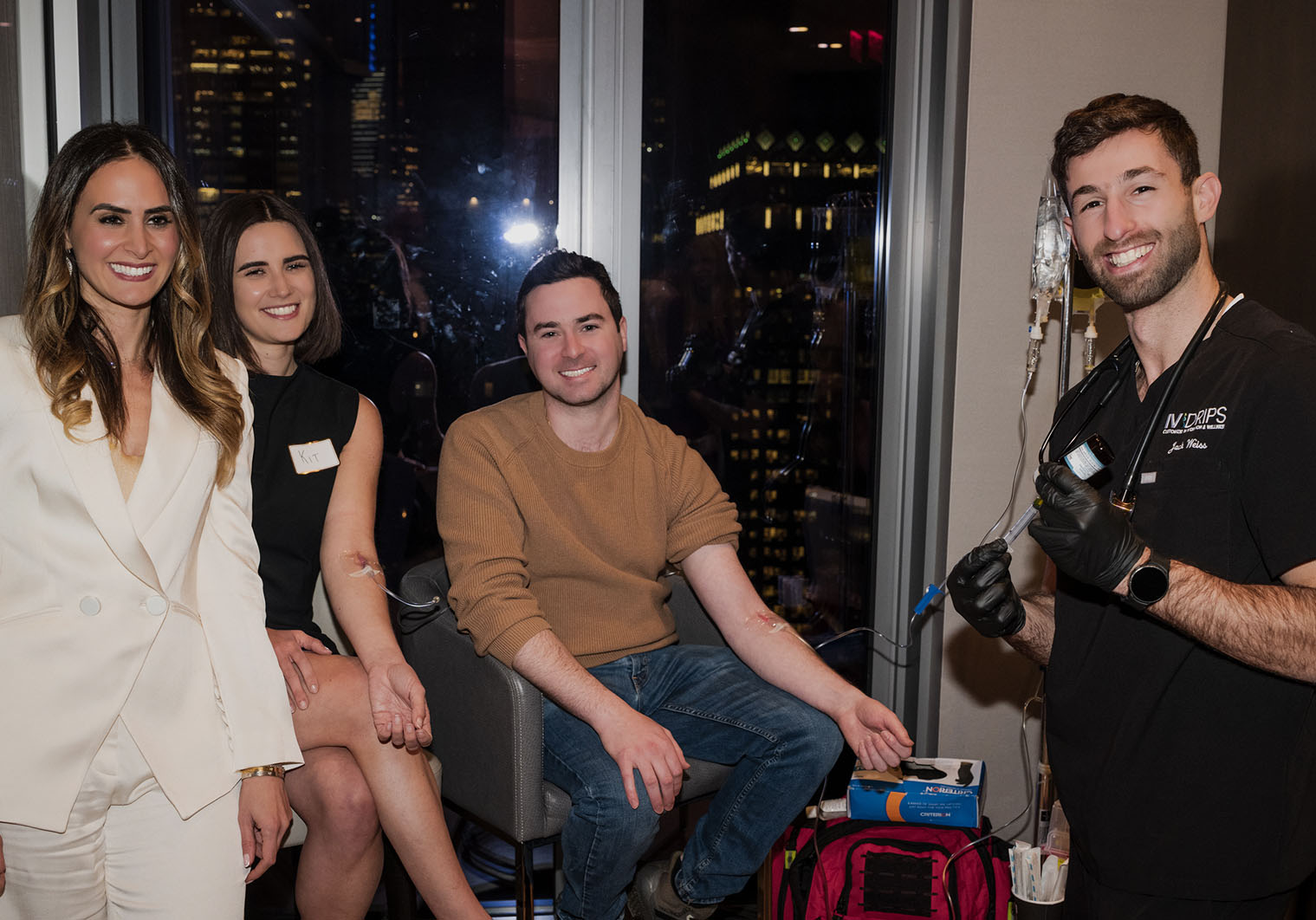 Four adults are indoors at night with city lights in the background. One man, wearing gloves, prepares an IV for another seated man, while two women stand and sit nearby, all smiling at the camera.