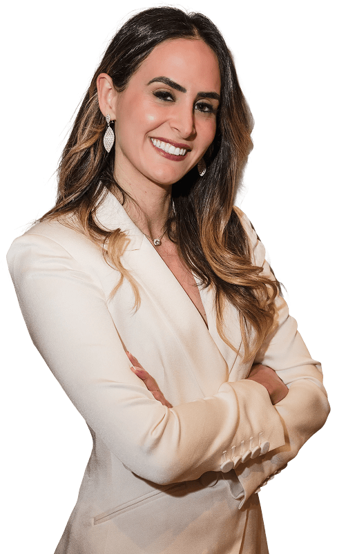 A woman with long, wavy brown hair wearing a white blazer stands with arms crossed, smiling confidently at the camera. She is wearing earrings and a necklace, with a neutral background.